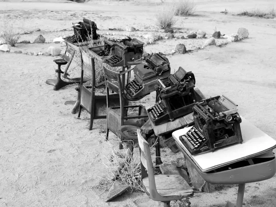 A black and white photo of a desert with old school desks, and typewriters on top of each one