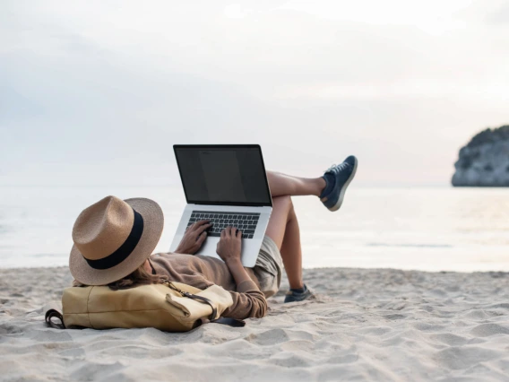 Image of a person on a beach relaxing but also typing on a laptop computer. 