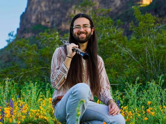 Portrait of Chris Zatarain with an oboe in a green field with yellow flowers and a mountain in the distance.