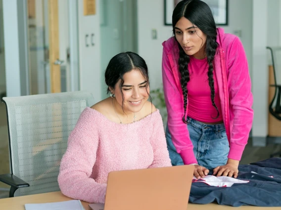 Two young women collaborate on a laptop and fabric spread on a desk in a bright office.
