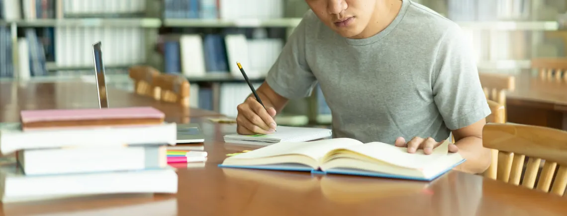 Person sitting at a desk writing in a notebook while reading from a book.