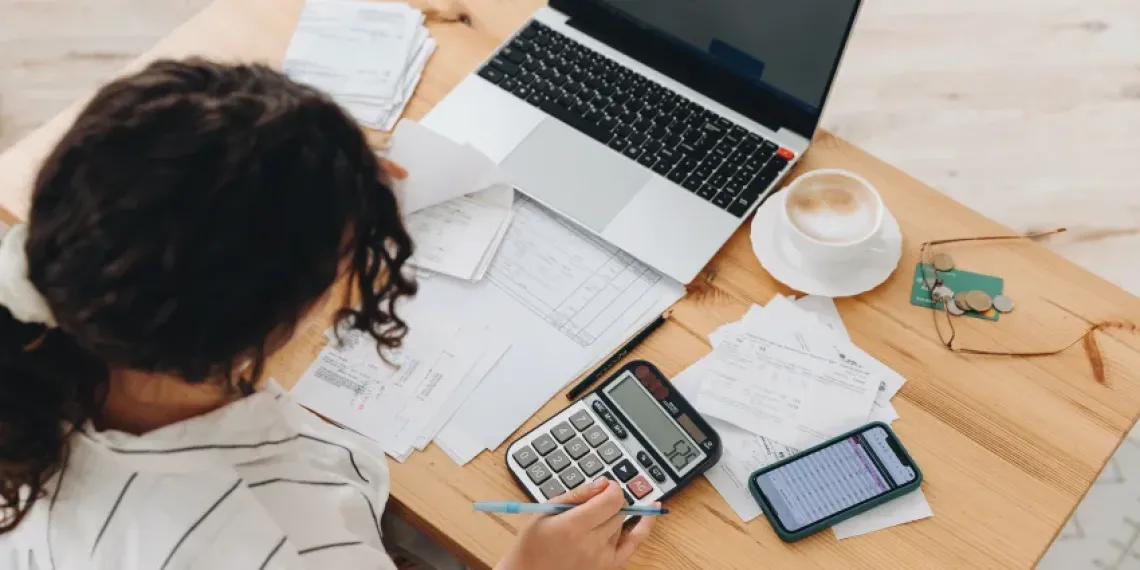 Person entering numbers on a calculator surrounded by financial documents, a laptop, and a smartphone. 