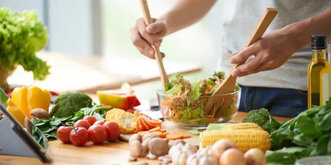 Hands holding wooden utensils mixing a salad on a table, surrounded by vegetables