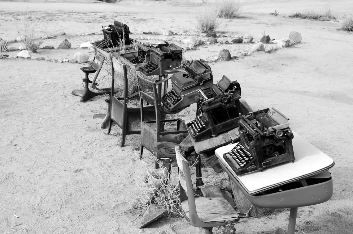 A black and white photo of a desert with old school desks, and typewriters on top of each one