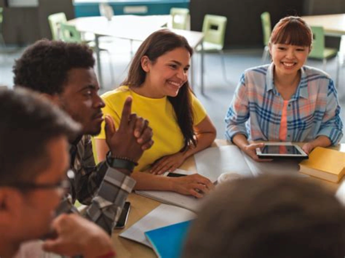 Diverse group of students collaborating around a table with books and a tablet.