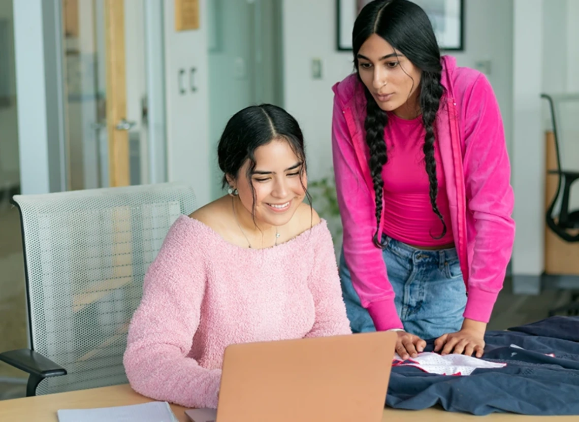 Two young women collaborate on a laptop and fabric spread on a desk in a bright office.