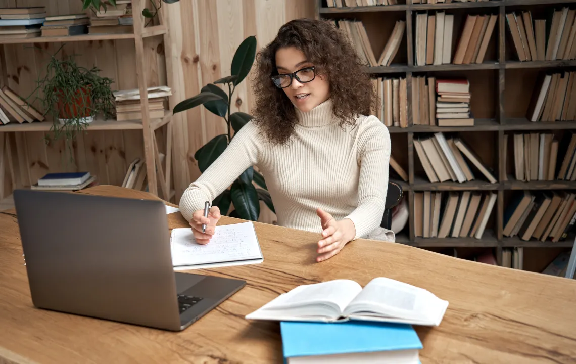 A woman in front of a bookshelf speaking to a laptop