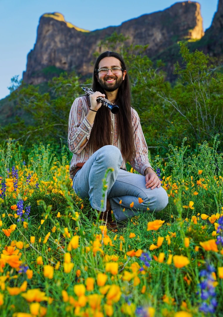 Portrait of Chris Zatarain with an oboe in a green field with yellow flowers and a mountain in the distance.
