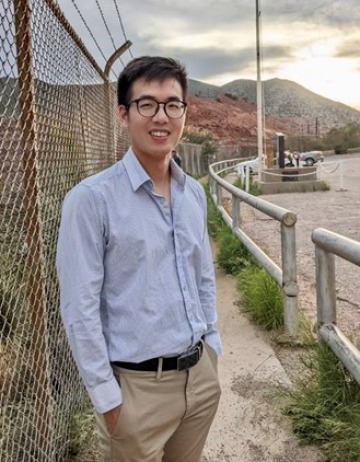 Zhixue Shu in a blue button-up shirt and khaki pants standing in front of rolling hills and mountains.