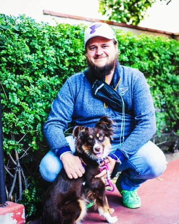 Josh in a cap and jacket crouching next to a brown dog with a pink leash