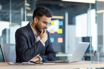 A man in a suit sitting in front of a laptop contemplating. 