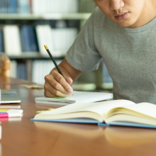 Person sitting at a desk writing in a notebook while reading from a book.