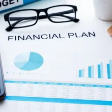 A document titled Financial Plan surrounded by a laptop, pen, glasses, calculator, and a binder.