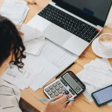 Person entering numbers on a calculator surrounded by financial documents, a laptop, and a smartphone. 