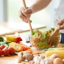 Hands holding wooden utensils mixing a salad on a table, surrounded by vegetables