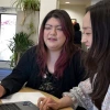 Two women sitting at a table looking at a computer. One woman is smiling and the other is speaking.