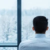 A man in a suit sitting in front of a window looking out onto a snowy landscape. 