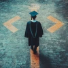 A person in Master's degree graduation regalia stands in the middle of a crossroad on a street. 