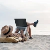 Image of a person on a beach relaxing but also typing on a laptop computer. 