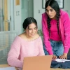 Two young women collaborate on a laptop and fabric spread on a desk in a bright office.