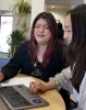 Two women sitting at a table looking at a computer. One woman is smiling and the other is speaking.